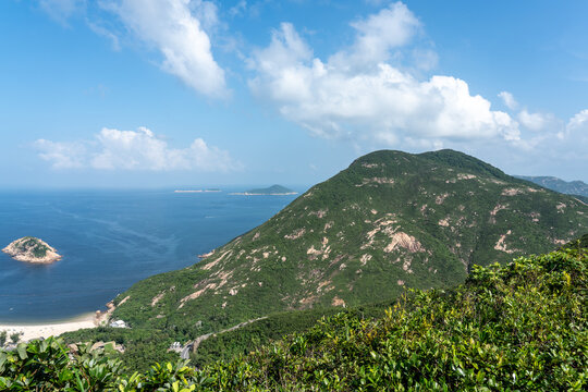 A breathtaking wide-angle panoramic view of lush green mountain slopes rolling down into a deep blue sea, featuring small rocky islands and a sandy beach.