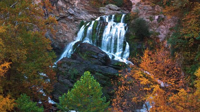 Turquoise waterfall cascades over granite boulders through peaceful woodland. Vibrant foliage and rocky formations create breathtaking seasonal landscape.