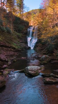 Waterfall autumn landscape with clear water tumbles from rocky boulders framed by orange and yellow forest trees.