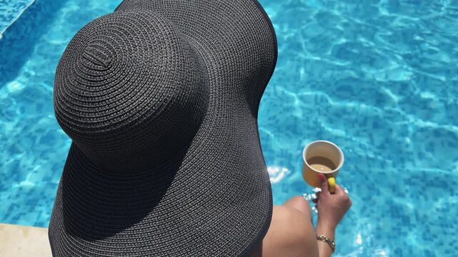 A woman wearing a black hat is sitting by a pool with a cup of coffee in her hand