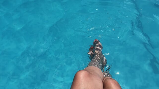 A woman is sitting in a pool with legs splashing. The hat is black and the water is blue