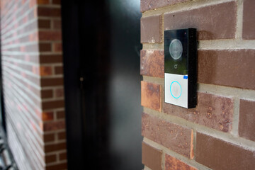 Smart doorbell camera mounted on red brick exterior wall beside residential front door in daytime,...