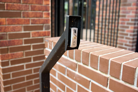 Metal keypad security access reader mounted on angled black post beside textured brick stairway wall in outdoor building entryway under bright daylight with soft visible shadows