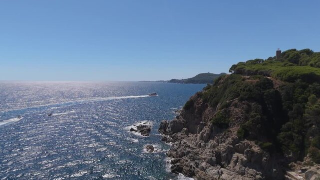 An aerial view of the Costa Brava coastline, Spain, taken with a drone, shows a boat sailing along the sea and coastal cliffs on a sunny day.