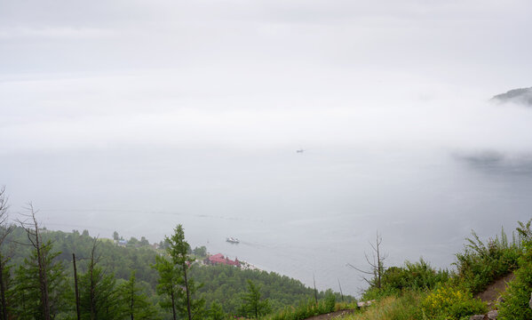 Elevated view of a foggy landscape with Lake Baikal and green forest shoreline, Russia. Small boats navigating on water under thick mist. Copy space for text