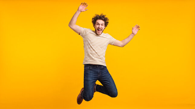 A man with curly hair and a beard jumping in the air with excitement, against a vibrant yellow background. He is wearing a light-colored t-shirt and dark jeans.