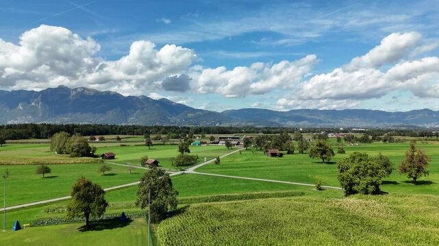 Aerial view of a wide green valley with farmland, cornfield and scattered barns below mountains in Austria