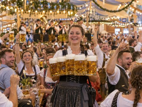 Lachende Bedienung im Dirndl tr&auml;gt mehrere gef&uuml;llte Bierkr&uuml;ge durch ein lebhaftes Festzelt mit Musik und feiernden G&auml;sten &ndash; bayerische Gastfreundschaft und ausgelassene Stimmung.
