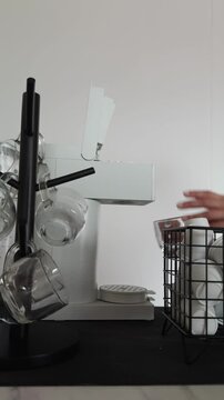 Hands taking a clear glass cup and placing it beneath the dispenser, then selecting a coffee capsule from a nearby storage basket