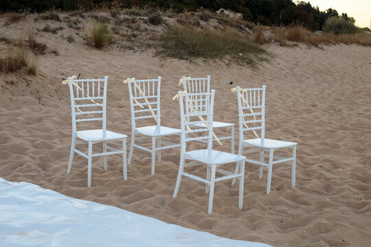 Row of white chiavari chairs decorated with bows standing on sandy beach dunes, arranged for an outdoor wedding ceremony