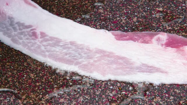 Bacon Slice for Scrambled Eggs. A strip of raw bacon is placed on the hot surface of the pan