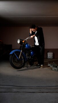 Man in work overalls feeling stressed and defeated while attempting to repair a vintage blue motorcycle with a toolbox and jerry can nearby in a dimly lit garage