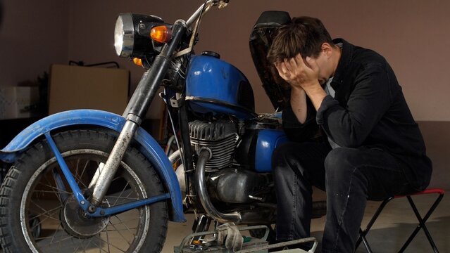 Young man sits in a home garage, hands covering his face in frustration and disappointment while unsuccessfully trying to repair his old broken motorcycle engine, diy struggle