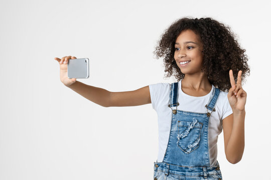 Cheerful young African girl using mobile phone, taking selfie and showing peace gesture isolated on white background, happy child, smartphone, social media, communication concept