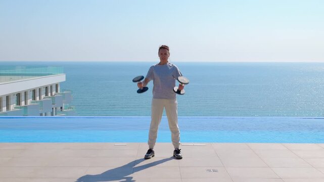 Woman doing bicep curls with dumbbells near an infinity pool. Athletic woman performing a bicep curl workout with dumbbells on a sunny day, standing by a infinity pool with a stunning ocean view