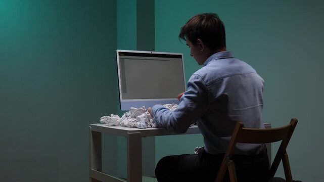 Frustrated man expressing stress and failure, emptying a full wastebasket of crumpled papers onto his computer desk, highlighting burnout and information overload