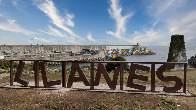 "LLANES" stand at a viewpoint overlooking the harbor pier and the colorful "Cubos de la Memoria" breakwater blocks, under a wide blue sky with wispy clouds, in Llanes, Asturias, Spain.