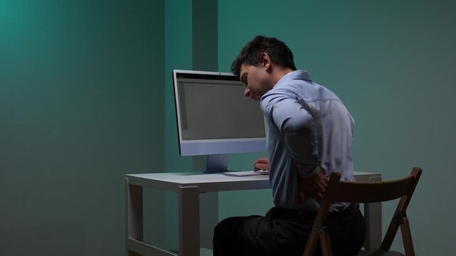 Man sitting at office desk clutching lower back in pain after long hours at computer, showing discomfort from sedentary work, poor posture and ergonomic strain