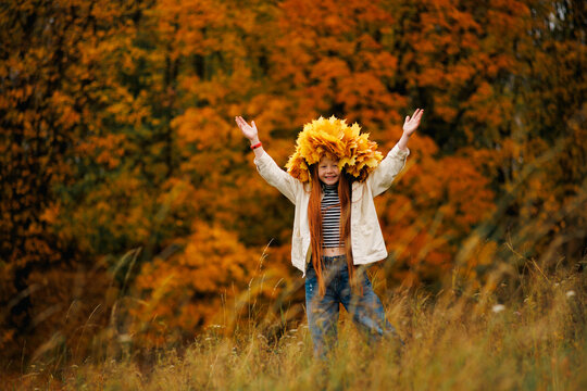 Joyful redhead girl in a maple leaf wreath raising her hands up on a grassy hill with blurred autumn trees background.