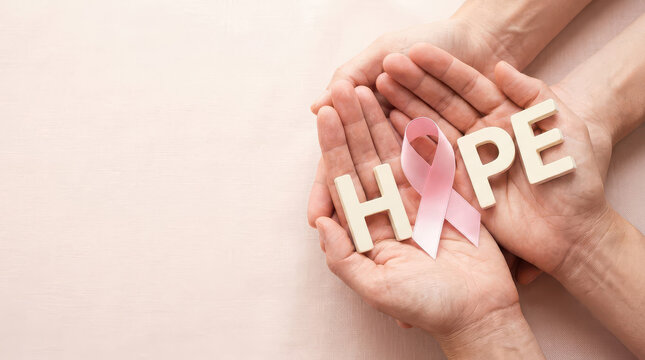 Two people stacking hands together holding wooden HOPE letters and pink ribbon on soft pink background, breast cancer awareness support concept for hope, unity and survivor encouragement