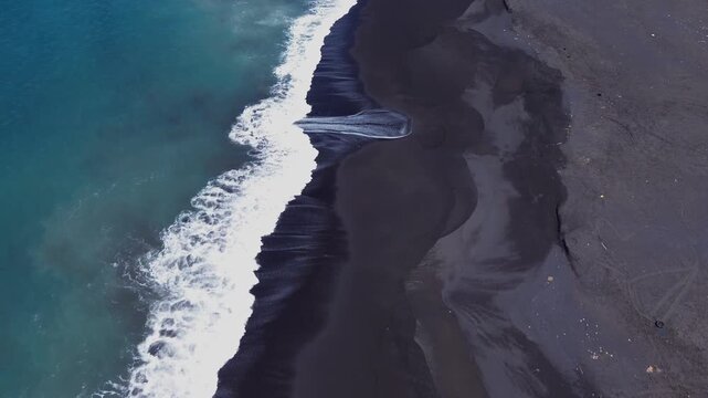 Birds eye top down aerial drone shot of white ocean wave foam crashing and flowing over black volcanic sand shoreline along Jumpai beach in Klungkung regency, Bali island, Indonesia.