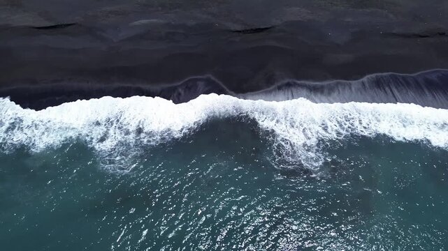 Birds eye top down aerial drone shot of white ocean wave foam crashing and flowing over black volcanic sand shoreline along Jumpai beach in Klungkung regency, Bali island, Indonesia.