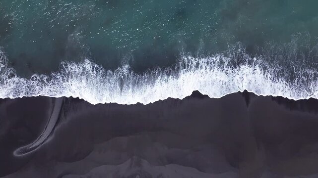 Birds eye top down aerial drone shot of white ocean wave foam crashing and flowing over black volcanic sand shoreline along Jumpai beach in Klungkung regency, Bali island, Indonesia.