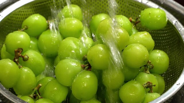 Macro view of fresh green grapes washed under flowing water in stainless steel colander in kitchen.