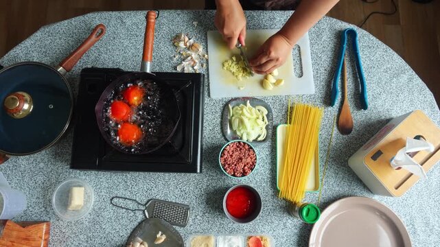 Top-down view of cook finely chopping garlic cloves surrounded by colorful pasta ingredients