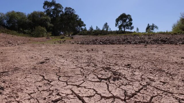 Low horizontal slider shot of cracked, parched soil beneath a cloudless blue sky in rural Portugal. Harsh sunlight, dramatic ground texture, and distant trees evoke extreme drought conditions.