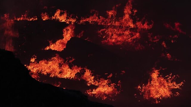 Dramatic nighttime wildfire rages through dense Amazon forest, sending waves of orange flames and thick smoke into the dark, chaotic air. Fiery inferno, devastation.