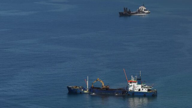 Drone shot captures a split hopper dredger operating near Madeira's serene coastline, with blue waters, clear skies, and another ship in the background under bright daylight.