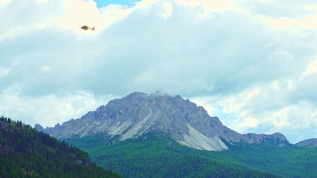 Yellow emergency rescue helicopter flying across dramatic mountain peaks and cloudy sky in the Italian Dolomites