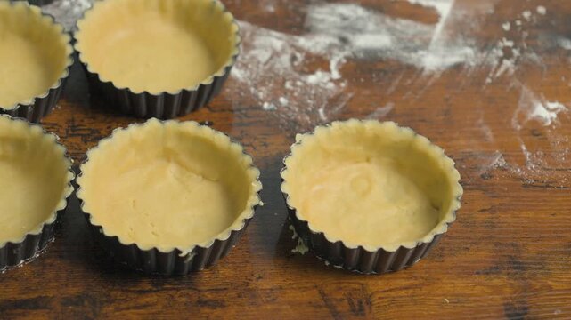 Pastry chef rolling out dough for tartlets on a floured wooden work surface, with several tart molds prepared for baking. Close-up of traditional pastry preparation in a bakery kitchen.