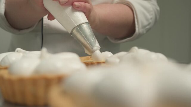 Pastry chef piping meringue onto lemon tartlets using a pastry bag, showcasing the preparation of a homemade dessert in a professional bakery setting. Close-up food preparation with shallow depth of f