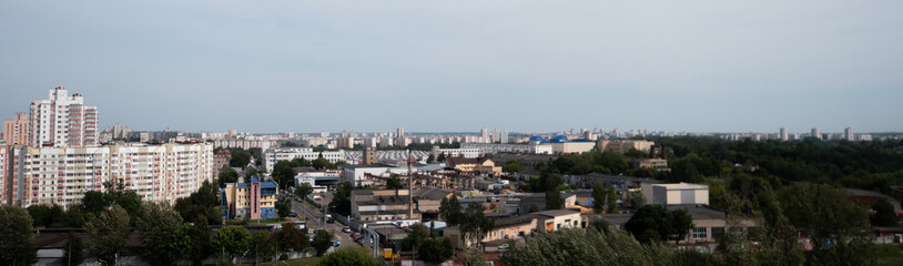 Obraz premium Wide urban skyline with apartment blocks, low-rise buildings, trees, and distant city horizon under soft cloudy sky, showing dense residential districts and calm summer atmosphere