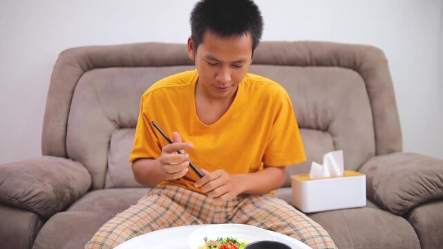 Young asian man eating delicious noodles with chopsticks on cozy sofa at home
