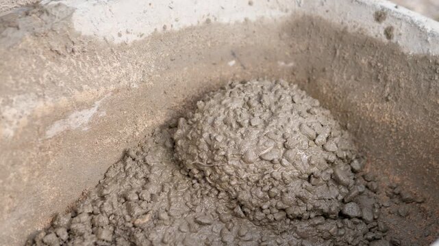 Wet gray concrete mix fills rough stained container in close-up, showing thick lumpy aggregate, dense viscous surface, muted earthy tones, soft diffuse light, and raw construction material texture