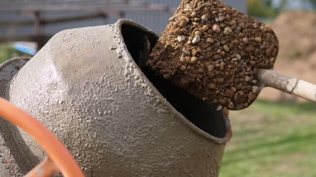 Close view of mud covered mixer drum or hopper shows rough wet texture, dark interior, and heavy wear outdoors beside blurred industrial structures, stacked material, and grassy ground in soft warm