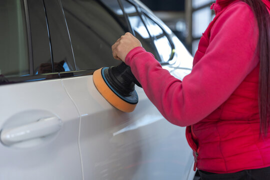 A woman works on polishing the side of a white car in an auto detailing shop. She focuses on her work.