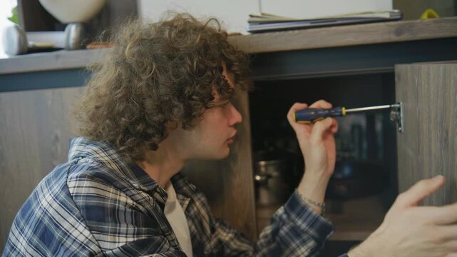 Young man works on fixing cabinet with screwdriver in home setup near kitchen in afternoon light
