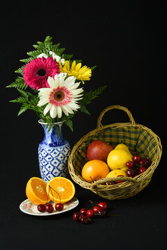 Naturaleza muerta con flores de gerbera coloridas, cesta con frutas variadas y naranjas cortadas sobre fondo negro.