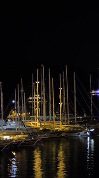 Vertical video, timelapse: moored yachts and gulets at night in calm harbor, reflecting lights and fast ripples, waves on the water surface - time lapse