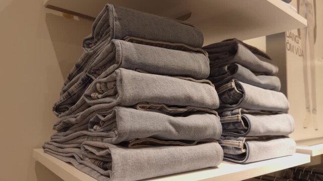 Stacks of neatly folded blue jeans displaying denim texture and material, creating a repetitive pattern on a clothing store shelf, representing fashion retail and apparel industry