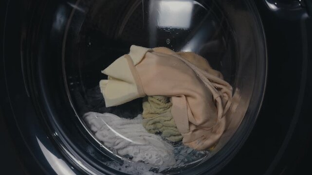 Washing machine drum spinning wet clothes and fabrics with soap suds and water droplets inside, close up of front loader action showing cleaning, motion and household laundry care