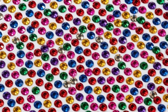 Assortment of shiny multicolored craft rhinestones arranged on white surface. Small circular gem stickers in red blue green yellow pink purple for decoration or art projects.