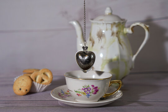 aza de t&eacute; vintage con infusor de coraz&oacute;n, galletas y tetera de porcelana al fondo.