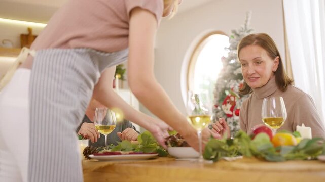 Senior parents and adult daughter enjoy Christmas dinner together at home. Happy family sharing festive holiday meal with roast chicken, salad and wine around dining table during seasonal celebration.