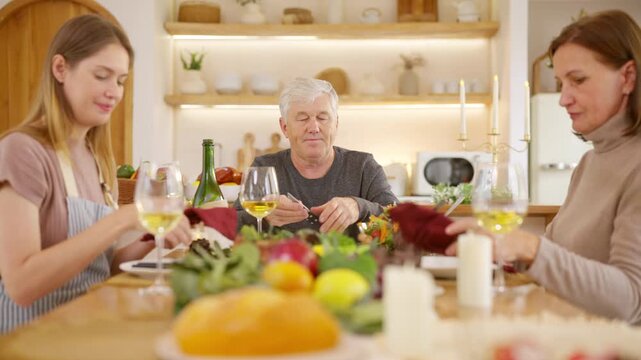 Senior parents and adult daughter enjoy Christmas dinner together at home. Happy family sharing festive holiday meal with roast chicken, salad and wine around dining table during seasonal celebration.