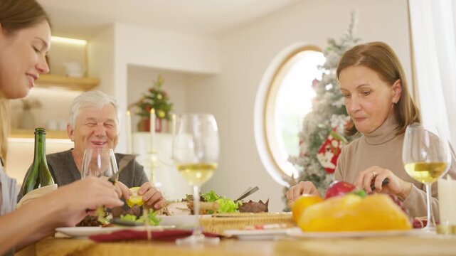 Senior parents and adult daughter enjoy Christmas dinner together at home. Happy family sharing festive holiday meal with roast chicken, salad and wine around dining table during seasonal celebration.